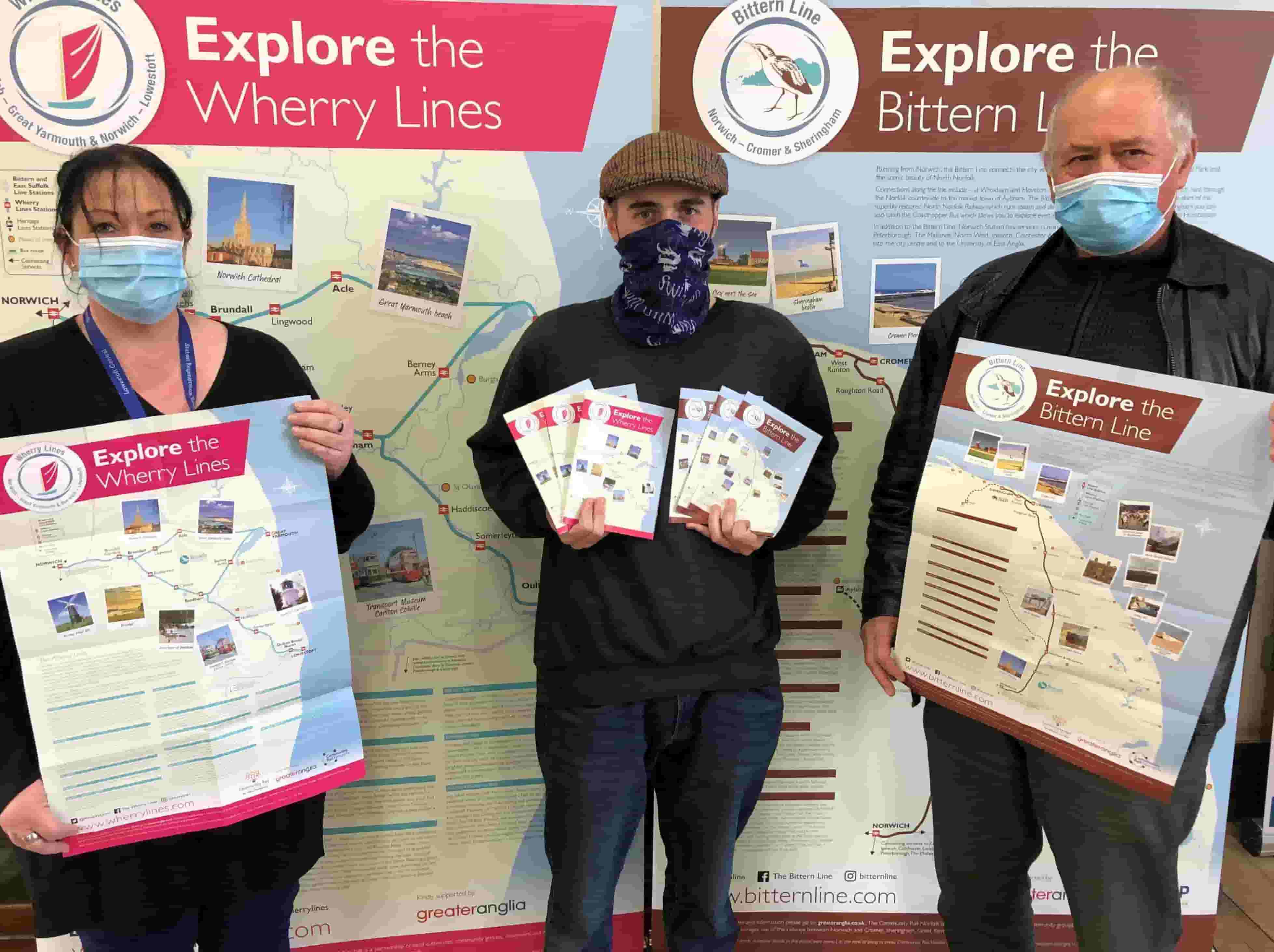 One woman and two men in face coverings holding 'explore the wherry lines' and 'explore the bitten line' posters 