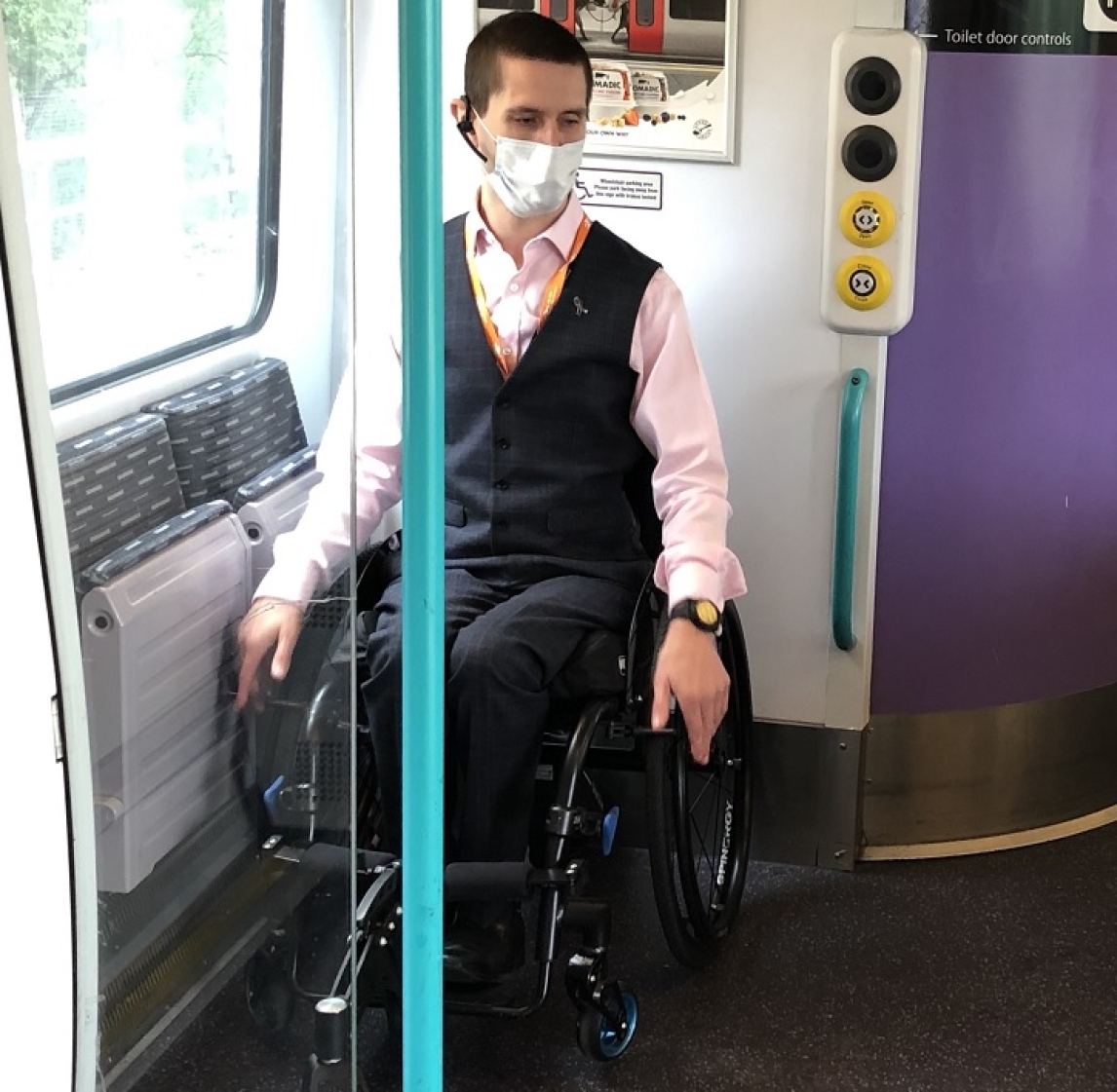 Train passenger in a wheelchair in a wheelchair space on a train, wearing a face covering.